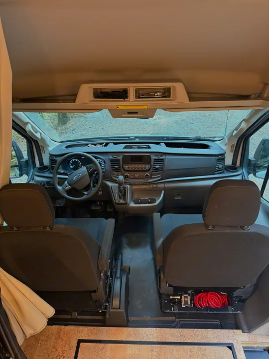 RV rental: Front cab view of a Ford van's dashboard, steering wheel, and seats. Under-seat storage with red hose visible.. Ford Transit 2023