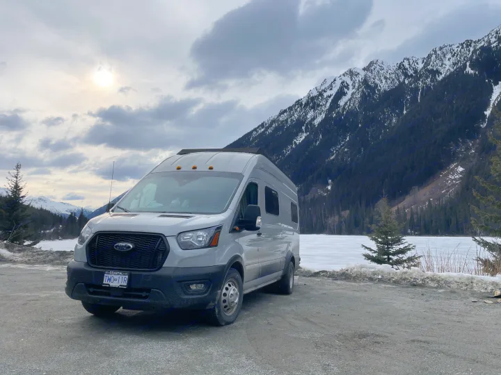 Grey Ford Transit RV rental parked by a frozen mountain lake, surrounded by snowy peaks and evergreen trees under a cloudy sky.. Ford Transit 2023