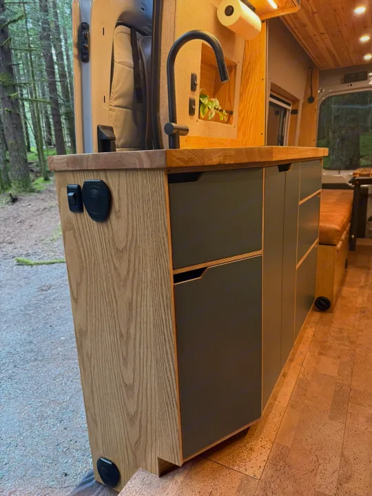 Inside a modern RV rental, a sleek kitchen with wood cabinets, dark drawers, sink, and faucet. A forest view is outside the door.. Ford Transit 2023