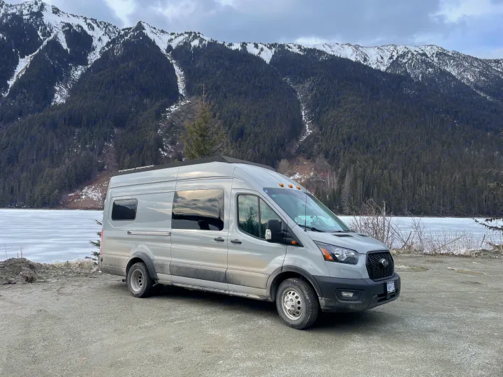 Light grey RV rental van parked by a frozen lake, with dense forest and snow-capped mountains in the background, perfect for nature travel.. Ford Transit 2023