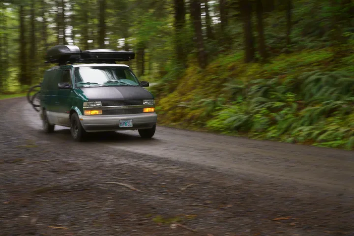 Panned shot of a green RV rental van with roof cargo & bike rack speeding down a gravel forest road.. Chevrolet Astro Van AWD 1998