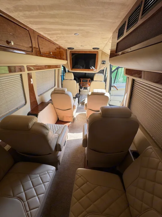 Step inside this luxurious RV rental: a spacious interior with multiple rows of comfortable, quilted tan leather seats, rich wood trim, and a built-in TV for premium travel comfort.. Mercedes-Benz Sprinter 2015