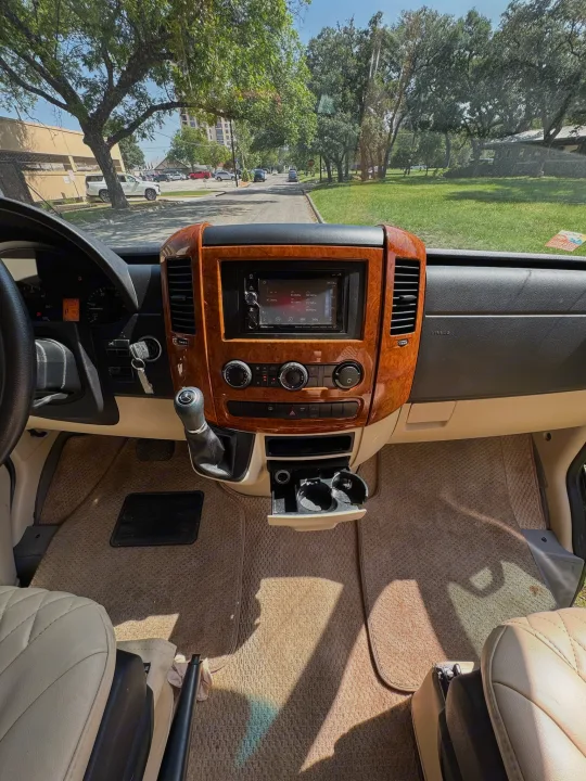 RV rental interior. Driver's view of dashboard with wood trim, screen, shifter, and sunny road ahead.. Mercedes-Benz Sprinter 2015