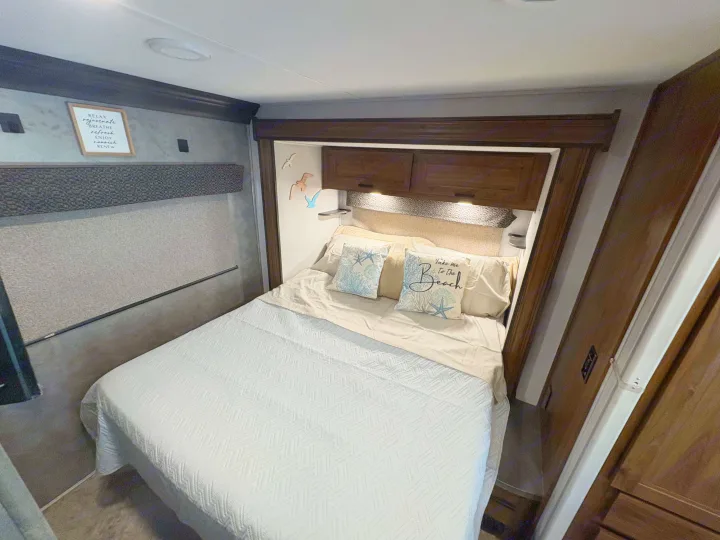 Relaxing RV rental bedroom featuring a comfy bed with coastal pillows & overhead cabinets. Enjoy a restful journey.. Forest River Sunseeker LE 2021