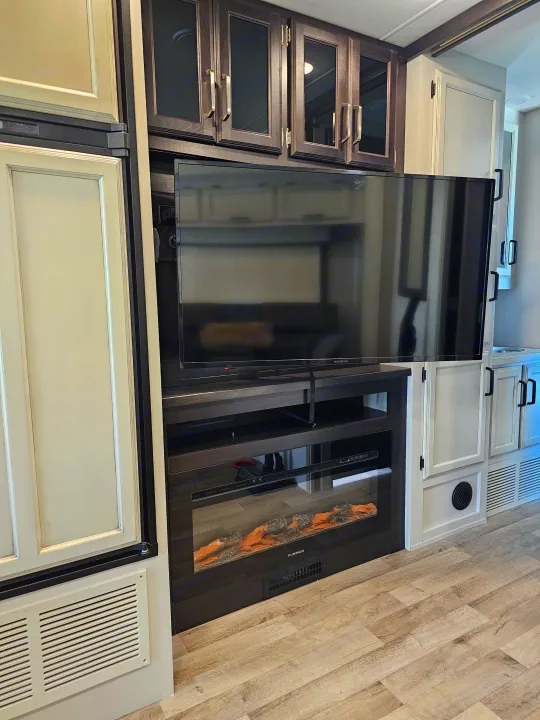 Inside a modern RV rental: a large TV & electric fireplace, flanked by a fridge & ample cabinetry.. Jayco Greyhawk 2023