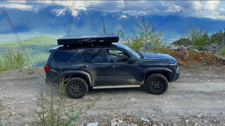 Dark SUV with rooftop tent on a mountain dirt road, against snowy peaks. Great for off-grid adventure beyond an RV rental.. Toyota 4Runner 2025