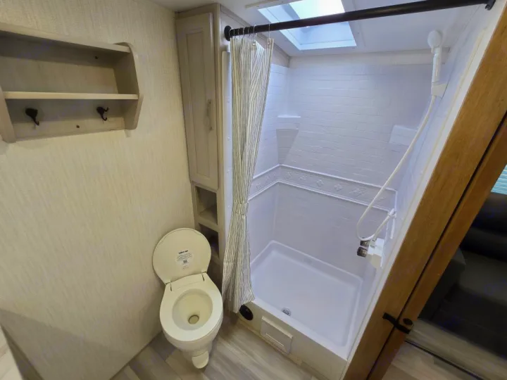 A clean RV rental bathroom showing a white toilet, shower with curtain, small wall shelf, and built-in cabinet.. Cruiser Embrace EL26RB 2024