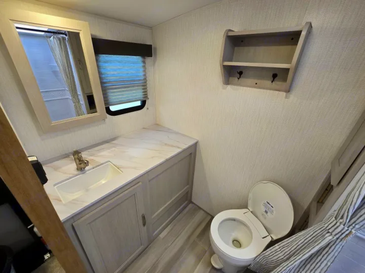 Inside an RV rental: a bright bathroom with light wood vanity, sink, mirror, toilet, and a wall shelf.. Cruiser Embrace EL26RB 2024
