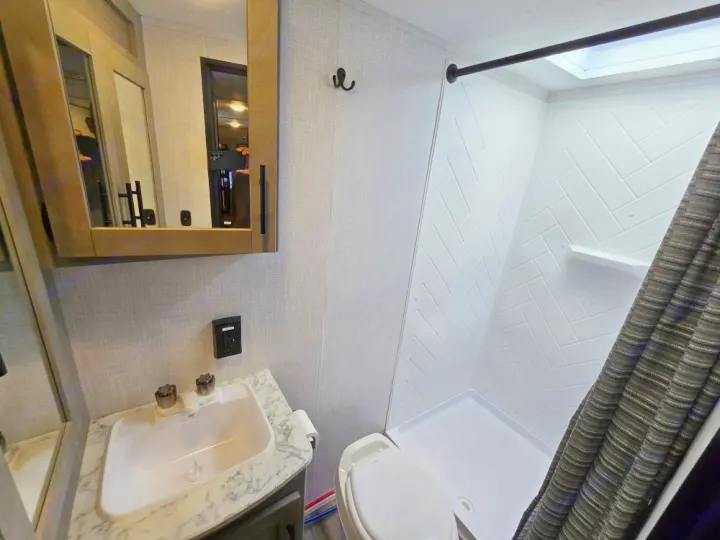 Inside a compact RV rental bathroom: a white sink on a marble counter, mirror cabinet, toilet, and white patterned shower.. Coleman 1855RB 2022