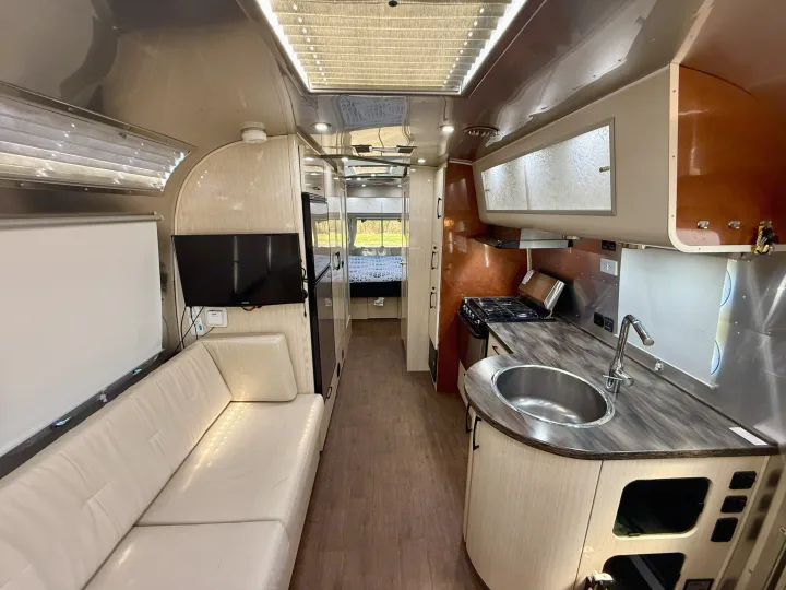 Spacious, modern RV rental interior: visible kitchen with sink/stove, white sofa, TV, and bed further back.. Airstream 27FBINT 2017