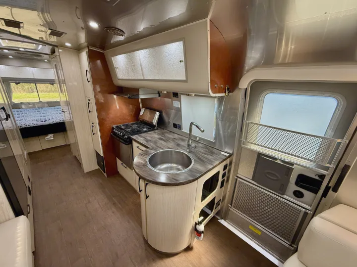 Modern RV rental interior: well-equipped kitchen with stove, sink, wood-look floor, and distant bed area.. Airstream 27FBINT 2017