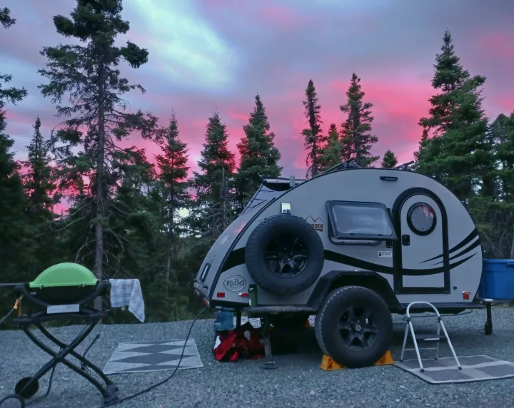 Vibrant pink sunset over a grey teardrop RV rental at a serene campsite, surrounded by tall pine trees. Ideal for nature lovers.. nuCamp T@G 2023