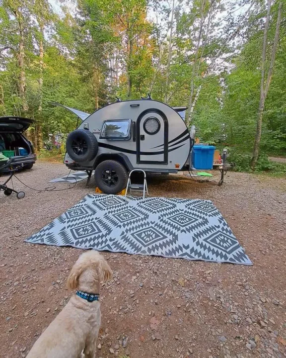 Teardrop RV rental at a wooded campsite, featuring a dog and an outdoor patterned rug.. nuCamp T@G 2023