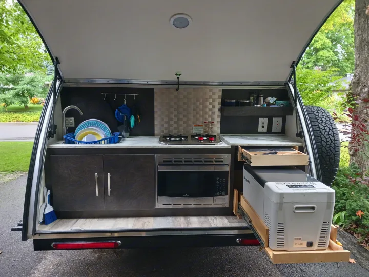Compact RV rental outdoor kitchen featuring a sink, stove, microwave, and a convenient pull-out fridge.. nuCamp T@G 2023