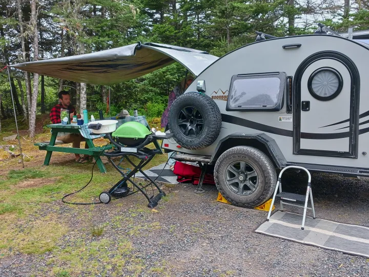 Man relaxes at a picnic table next to a grey teardrop RV rental setup with a grill and awning in a wooded campsite.. nuCamp T@G 2023