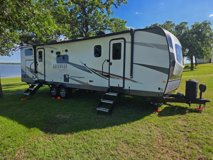 This RV rental, a white Rockwood travel trailer, is set up by a serene lake with green grass and trees under a blue sky.. Forest River Rockwood Ultra Lite 2022