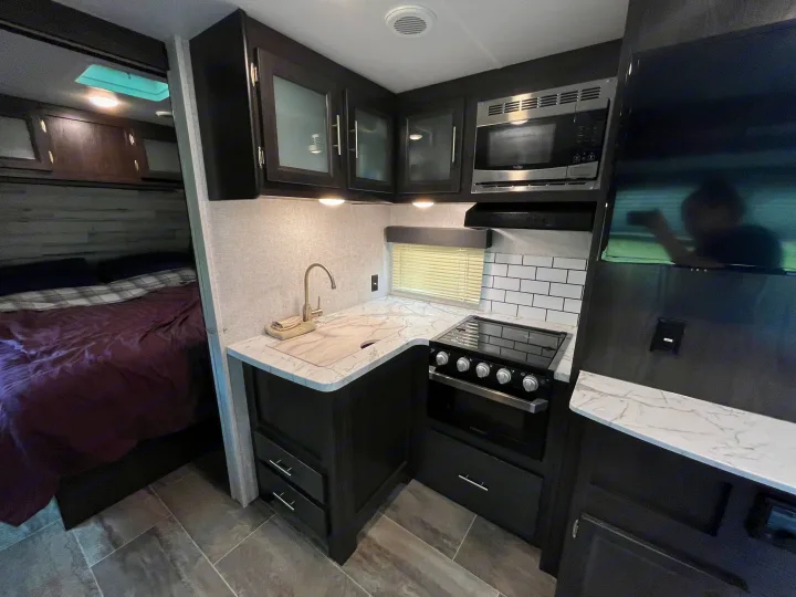 Modern RV rental interior: well-equipped kitchen with dark cabinetry, white counters, microwave, stove, and a cozy bedroom.. Dutchmen Kodiak Ultra Lite 2020