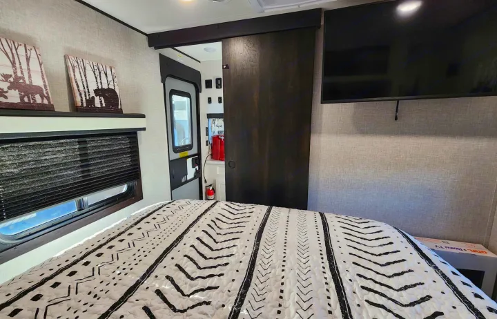 This modern RV rental bedroom features a cozy bed with a graphic comforter, wall art, a TV, and a dark wood sliding door.. Jayco Jay Flight 2024