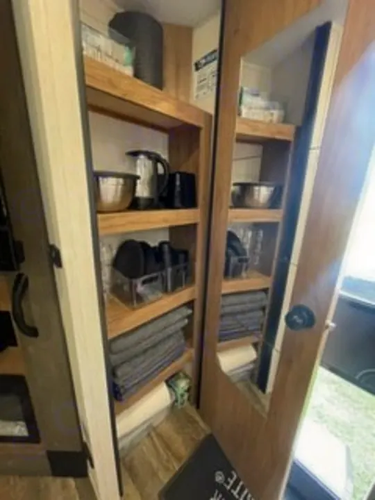 RV rental interior: An organized pantry with shelves of kitchenware and towels. A mirrored cabinet door is visible on the right.. Forest River Campsite Reserve 26CJ 2025
