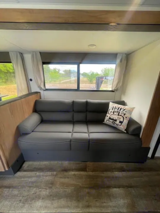 RV rental living space showing a dark sofa with a 'Happy Camper' pillow, large windows, and wood flooring.. Forest River Campsite Reserve 26CJ 2025
