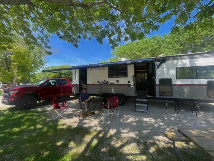 Relaxing at an RV rental campsite. A man and his dog enjoy the shade next to a red pickup truck and travel trailer.. Forest River Campsite Reserve 26CJ 2025