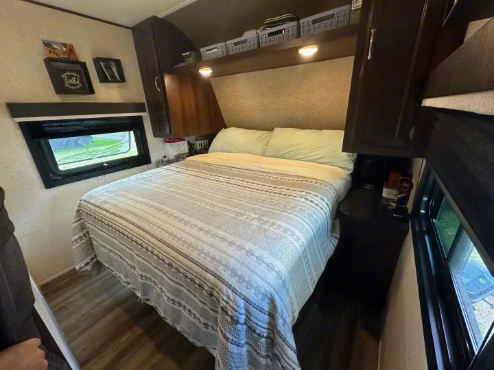 Interior of a comfortable RV rental bedroom, featuring a patterned bed, overhead storage, and a window.. Jayco Jay Flight 2018