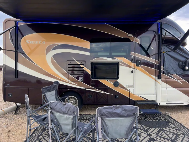 Relaxing at an RV rental site: a motorhome with blue lit awning, outdoor TV, and camping chairs on a rug.. Thor Siesta 2017