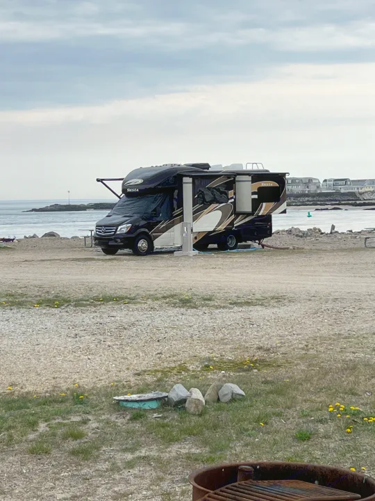 A sleek black and tan Siesta RV rental is set up at a seaside campsite, offering ocean views under a cloudy sky.. Thor Siesta 2017