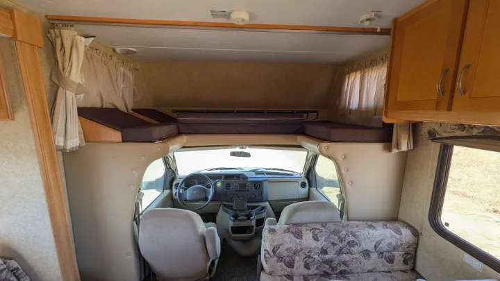 Inside a light-colored RV rental, featuring the driver's cabin, overhead bunk bed, and patterned sofa.. Forest River Sunseeker 2010