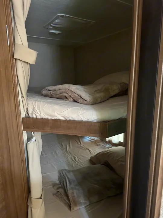 View of two compact bunk beds inside an RV rental, neatly made with light bedding and blankets. Curtains are on the left.. Gulf Stream Conquest 2017
