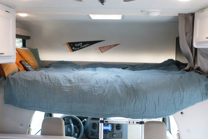 A cozy overhead bed in an RV rental, featuring blue bedding, decorative pillows, and a "Happy Camper" pennant.. Thor Motor Coach Eddie Bauer 21EBF 2025
