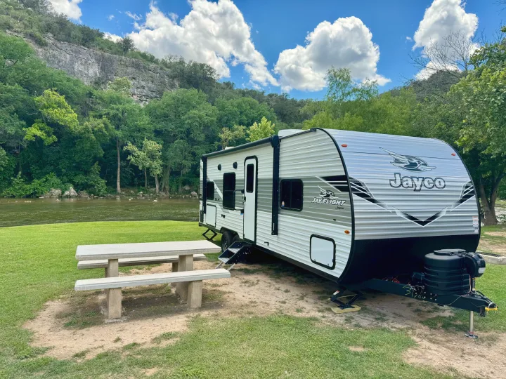 Riverside **rv rental** campsite: a Jayco travel trailer, picnic table, green grass, river, trees, and rock cliffs under blue sky.. Jayco 260BH 2025