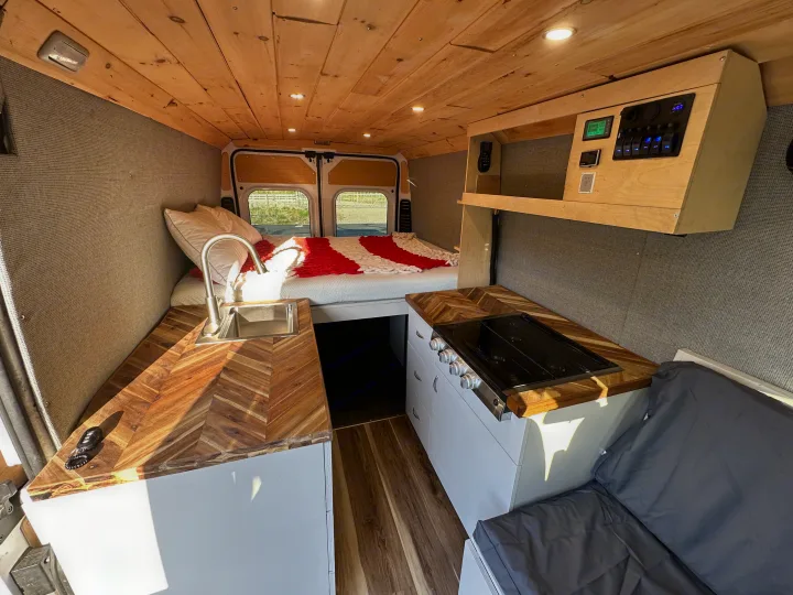 RV rental interior: wood-paneled ceiling, cozy bed, and compact kitchen with herringbone counters, sink, and stove. Control panel visible.. Dodge Promaster 3500 2020