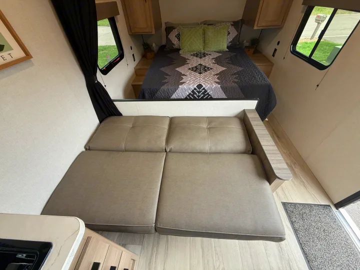 A cozy RV rental interior, featuring a bed with a geometric comforter, green pillows, and a gray seating area.. Keystone RV 23B 2025