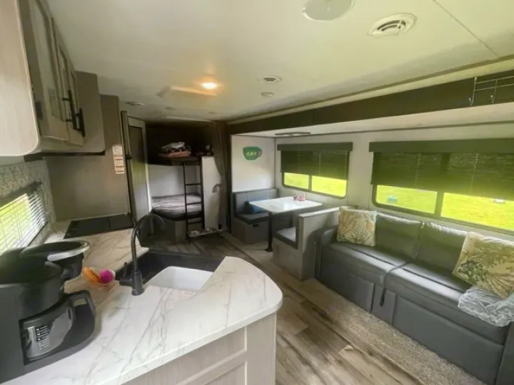 Inside a modern RV rental, showing a kitchen with sink and coffee maker, dining booth, comfy sofa, and bunk beds.. Coleman Lantern 2022
