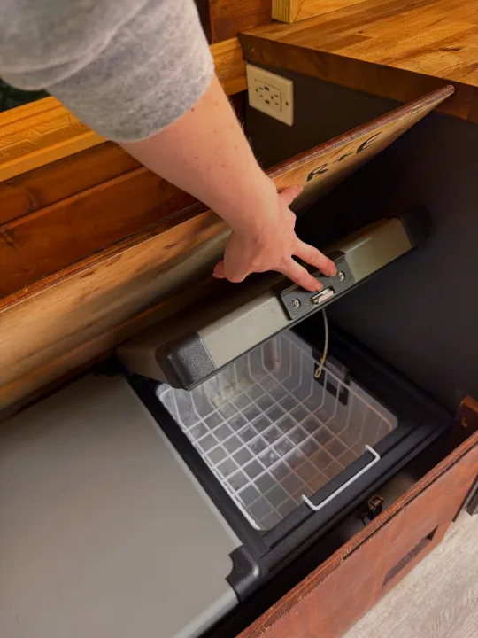 Hand opening a compact refrigerator/freezer with a basket, built into custom wooden cabinetry for an RV rental.. Mercedes Sprinter 2008