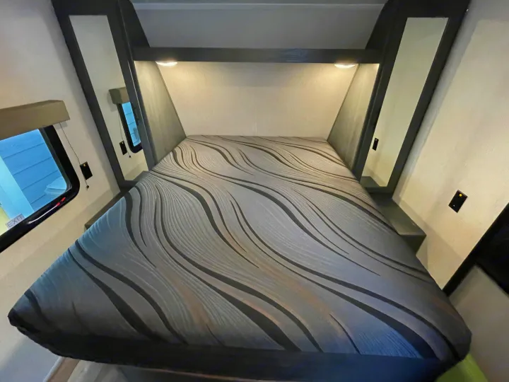 Comfortable RV rental bedroom with a grey wavy mattress, overhead lights, and side windows.. Dutchmen 23RKCWE 2024