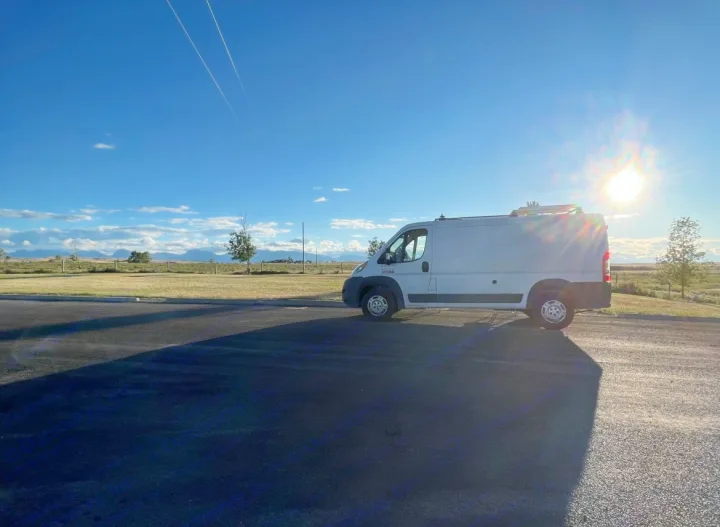 A white cargo van, perfect as an RV rental, parked on asphalt. Bright sun, distant mountains, & open fields under a blue sky.. RAM Promaster 2017