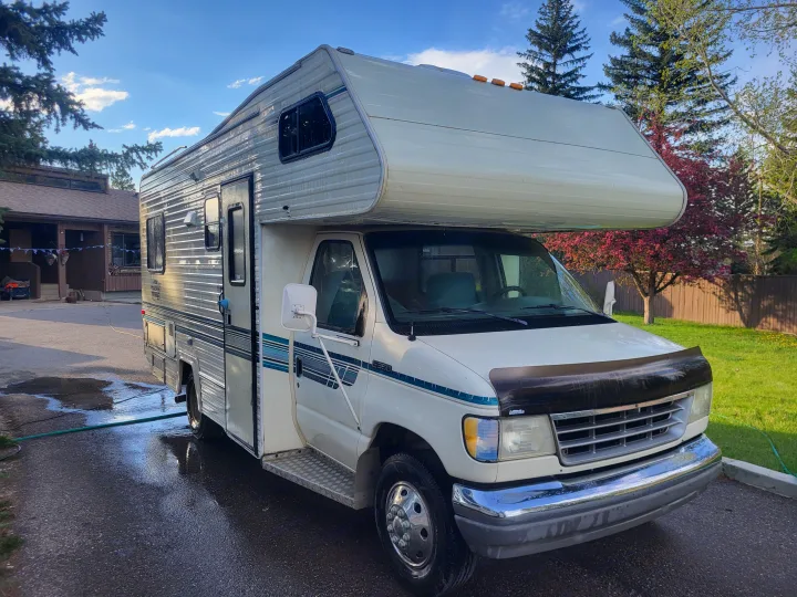 A white Class C RV, possibly an RV rental, is parked on wet asphalt in a residential setting.. Ford Econoline 1992