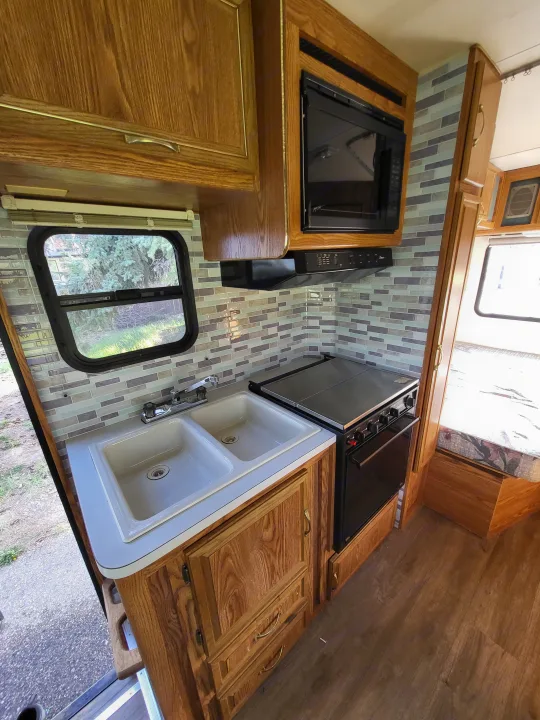 Clean RV rental kitchen with dual sink, stove, microwave, and modern mosaic backsplash. Warm wood cabinets line the space.. Ford Econoline 1992