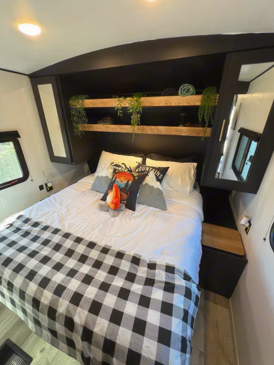 Cozy RV rental bedroom features buffalo plaid bedding, mountain-themed pillows, and overhead storage with shelves.. Keystone RV Springdale 2023
