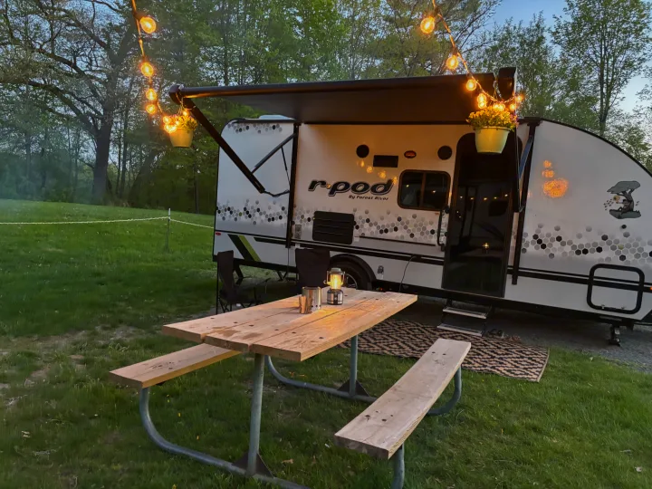 An RV rental parked at a peaceful campsite, featuring a wooden picnic table with a lantern, glowing string lights, and chairs for camping comfort.. Forest River R-Pod 2022