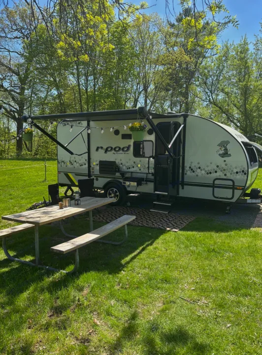 An r-pod RV rental at a sunny campsite with a picnic table and green grass, surrounded by trees.. Forest River R-Pod 2022