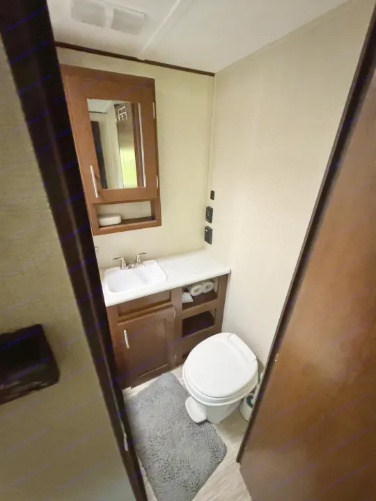 Compact RV rental bathroom with a sink, toilet, dark wood vanity, medicine cabinet, and a gray rug. Efficient space.. Forest River Avenger ATI 2018