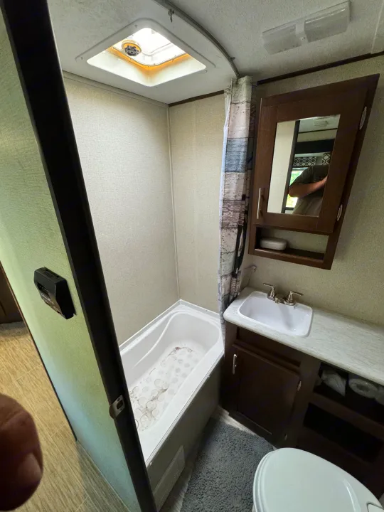 The bathroom in an RV rental, showing a compact tub/shower, vanity with sink, and toilet. A skylight brightens the space.. Forest River Avenger ATI 2018