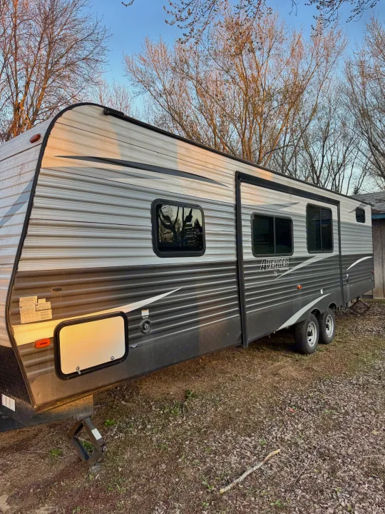 This gray & white Avenger travel trailer RV rental is parked outdoors amidst early spring trees, ready for your next trip.. Forest River Avenger ATI 2018