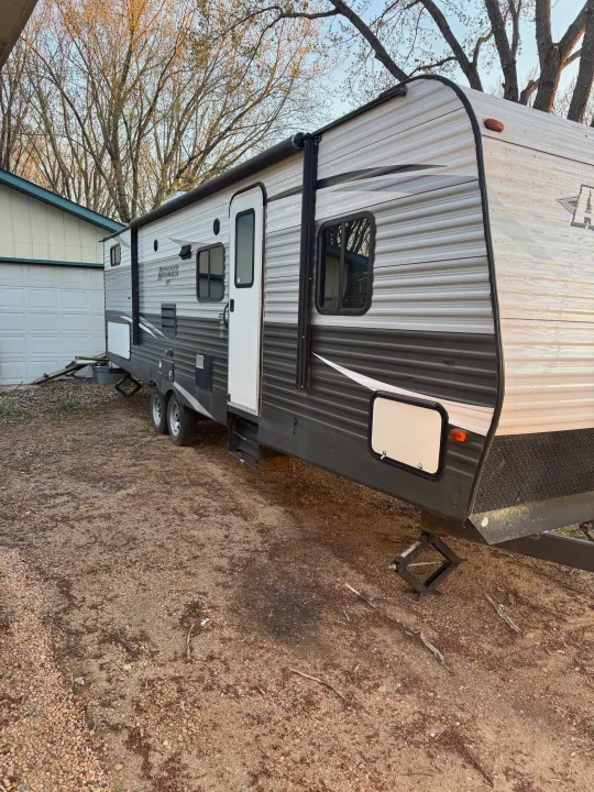 A grey travel trailer RV rental, parked on a gravel drive with leveling jacks deployed next to a garage.. Forest River Avenger ATI 2018
