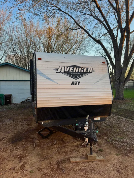 White Avenger travel trailer, an RV rental option, parked on a dirt lot with a house and trees in the background.. Forest River Avenger ATI 2018