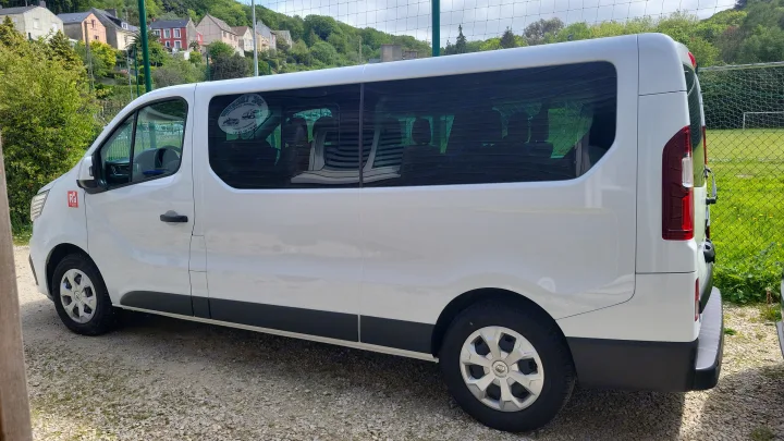White passenger van, ideal for RV rental, parked on gravel near a sports field. Houses dot the hillside.. RENAULT TRAFIC 2024