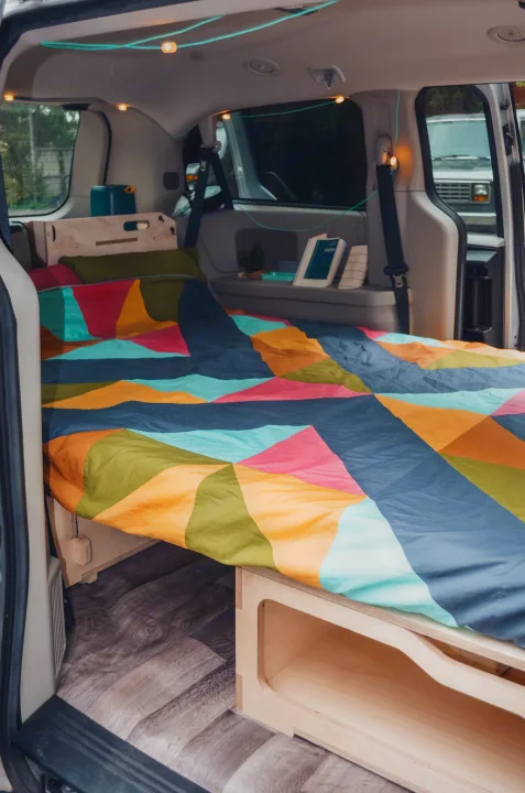 Colorful bed and clever storage in an RV rental campervan interior, enhanced by cozy string lights, ideal for mobile living.. Chrysler Grand Caravan 2025